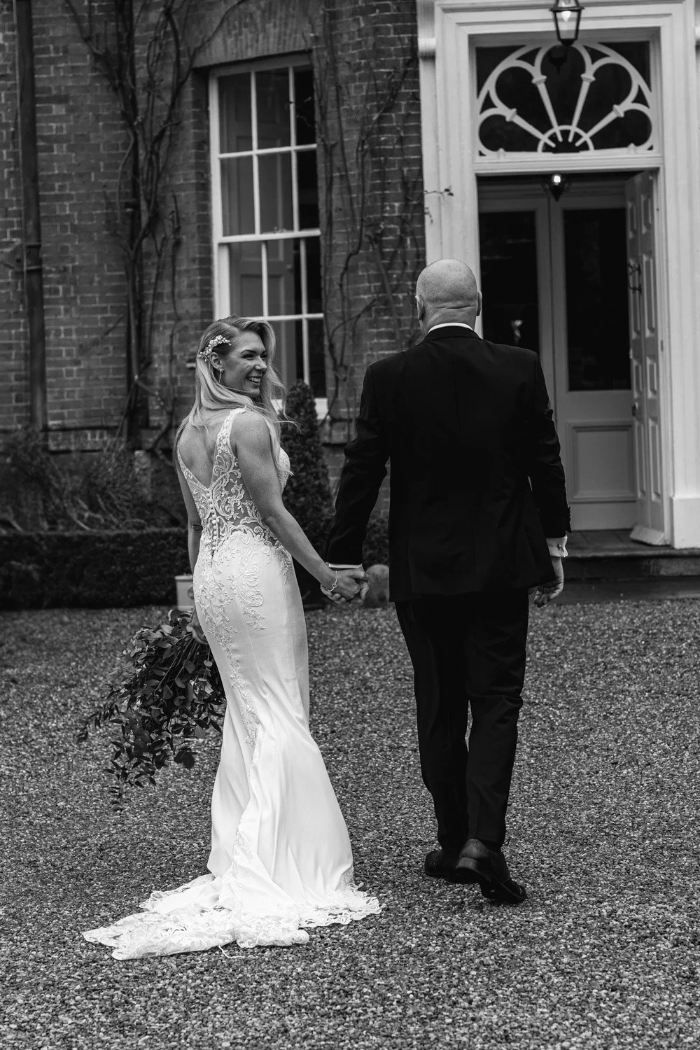 A bride and groom holding hands outside a building, with the bride smiling and looking back, in black and white.