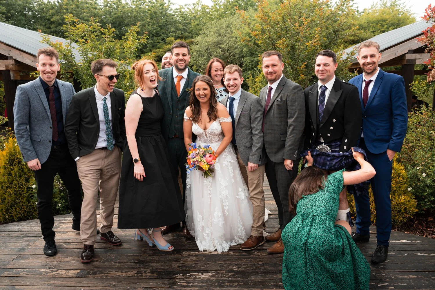 A group of people at a wedding, with the bride in a white dress holding a bouquet, surrounded by friends and family smiling and laughing outdoors in a garden setting.