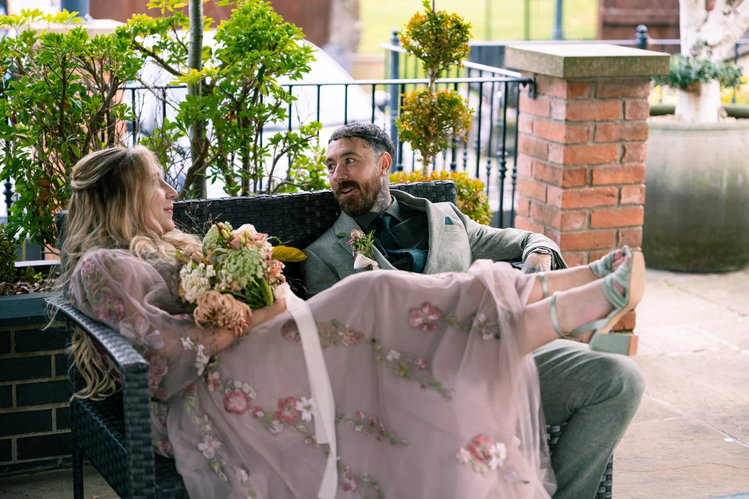 A couple sitting on a black wicker bench outside, with the woman in a pink floral dress holding a bouquet of flowers and the man in a grey suit with a teal tie, engaged in conversation, surrounded by potted plants and brick architecture.