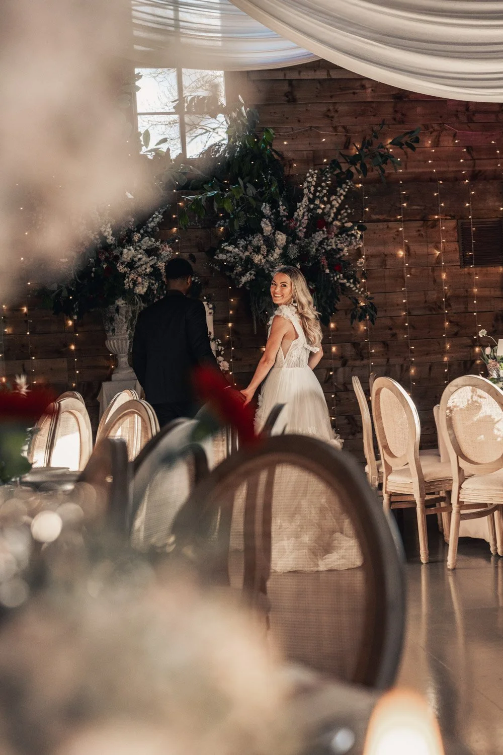 A bride and groom holding hands and smiling at each other at their wedding reception, decorated with flowers, fairy lights, and wooden walls, with empty chairs around them.