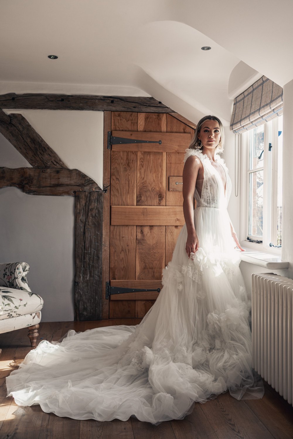 Bride in a wedding dress standing by a window in a rustic room with exposed wooden beams and a floral armchair.