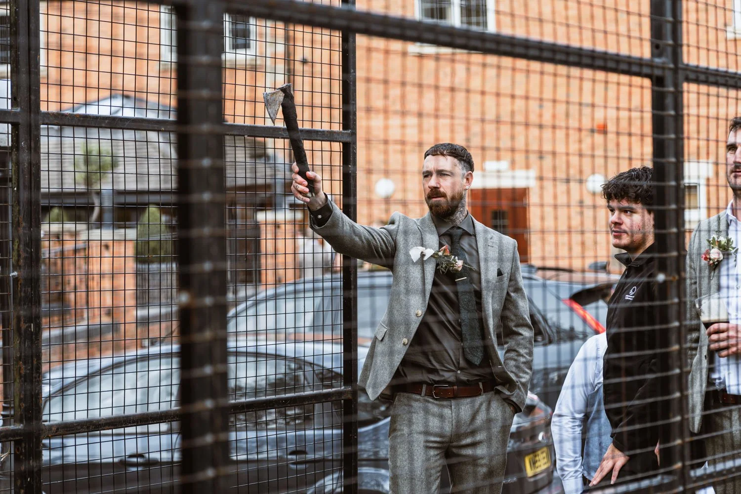 Three men in suits standing outside behind a metal fence. One man is holding an axe and appears to be about to chop the fence.