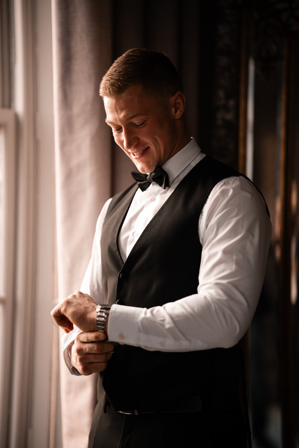 A man dressed in formal wear, adjusting his watch, smiling, standing near a window with curtains.