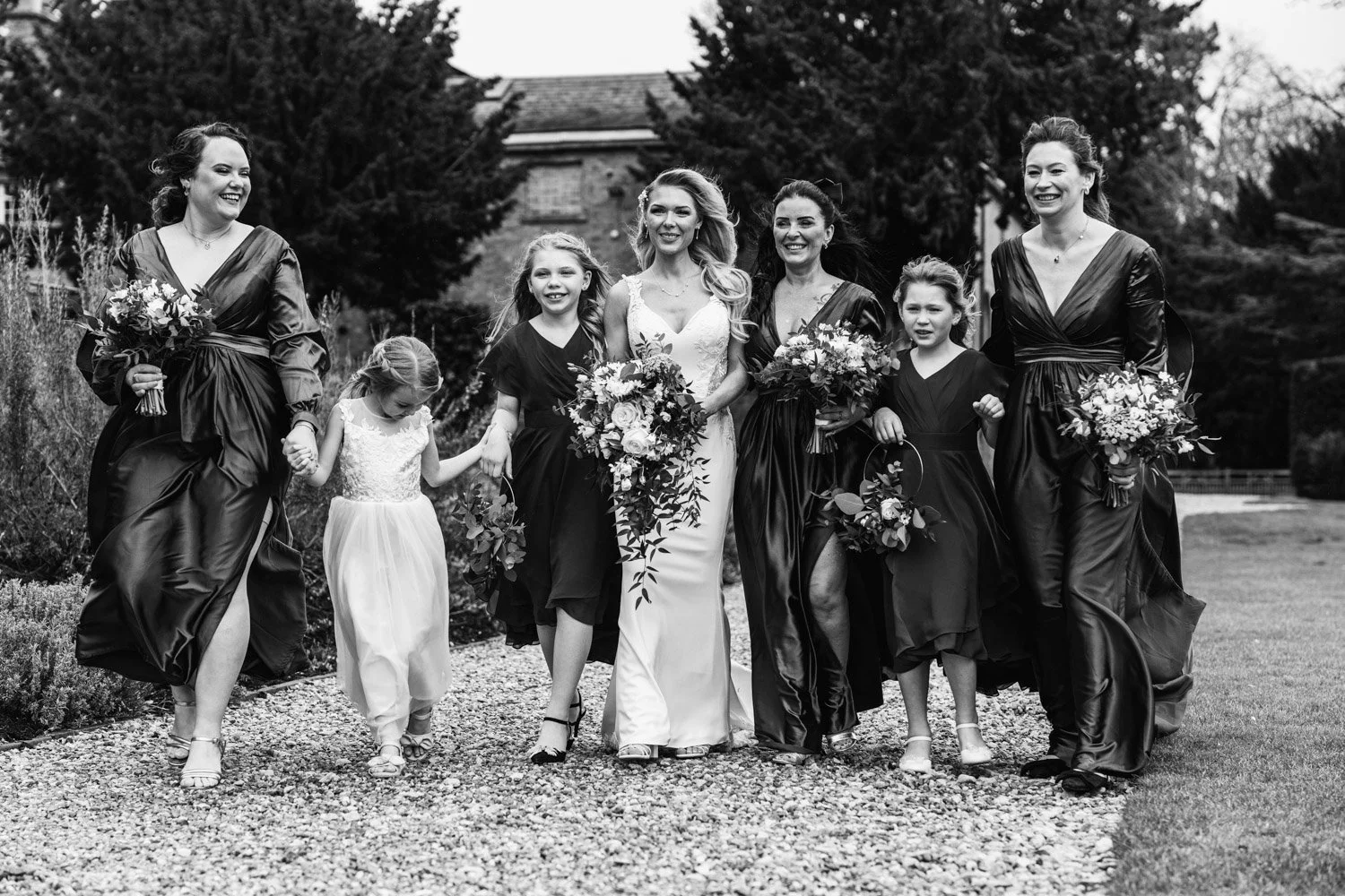 Black and white photo of women and young girls dressed in formal attire walking outdoors, holding bouquets of flowers, with trees and a building in the background.