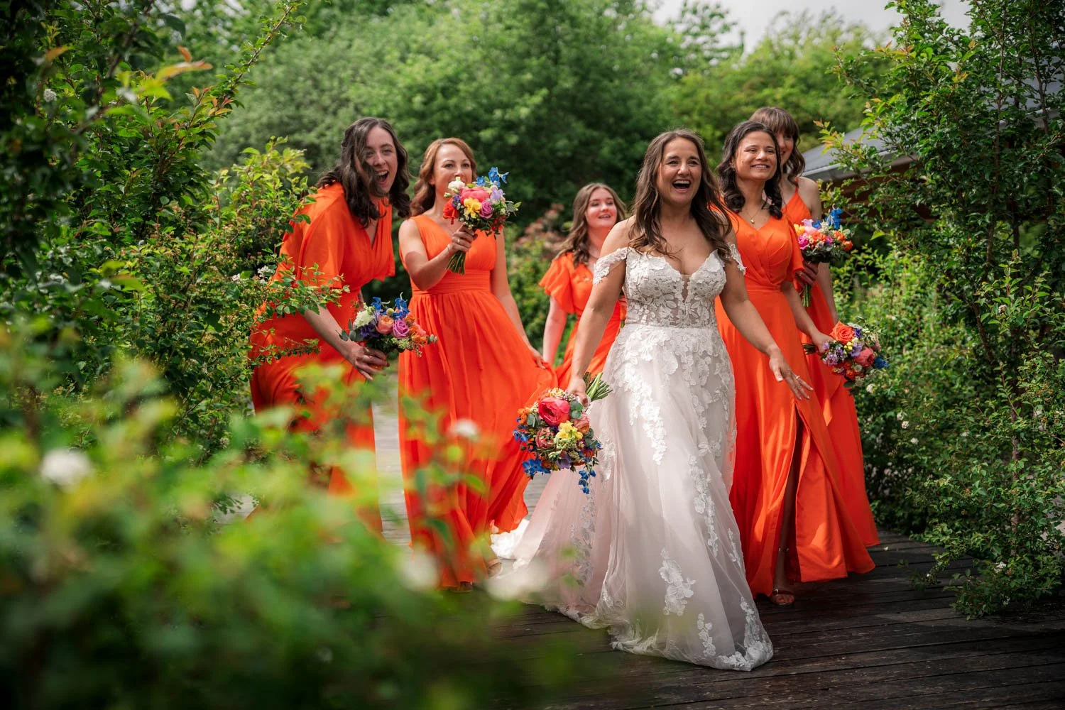 A bride in a white lace wedding dress walking with five bridesmaids in bright orange dresses, holding colorful bouquets in a lush outdoor garden setting.