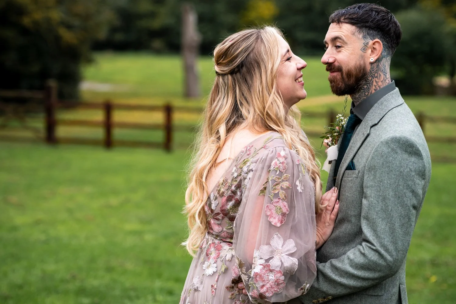 A smiling couple standing close together outdoors, with the woman in a floral dress and the man in a gray suit, facing each other affectionately in a green park.