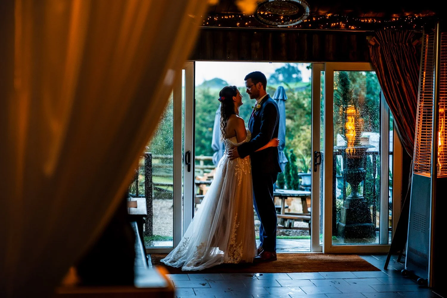 Bride and groom sharing a kiss inside near open sliding glass doors, with rain on the outside, surrounded by greenery, dim interior lighting, and wedding decor.