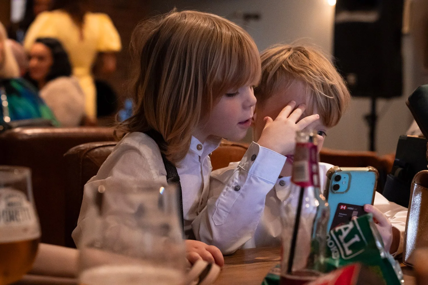 Two young children with long hair sitting at a table in a restaurant, looking at a smartphone together.