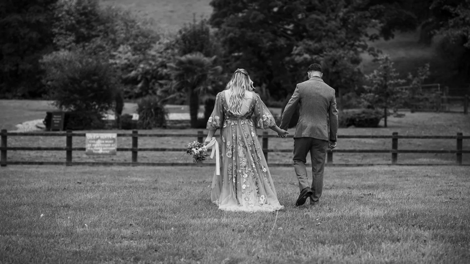 A couple walking hand in hand on a grassy field, with trees and a fence in the background. The woman is in a floral, flowing dress holding a bouquet; the man is in a suit.