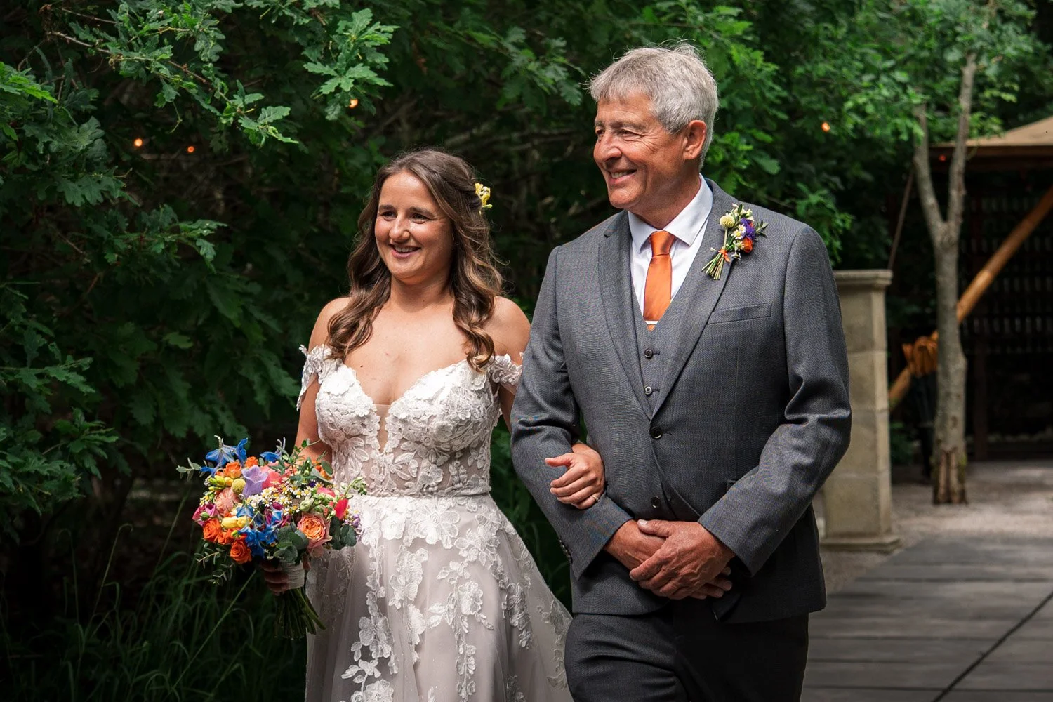 A bride in a lace wedding dress holding a colorful bouquet walks arm-in-arm with an older man in a gray suit, outdoors surrounded by green foliage.