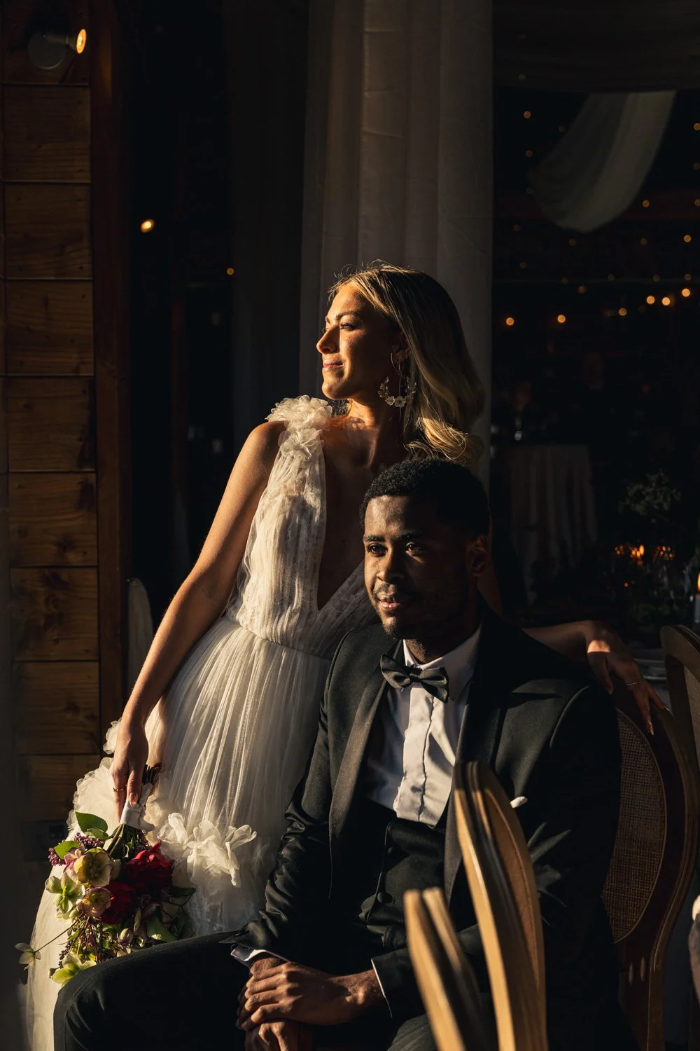 A woman in a white wedding dress standing beside a seated man in a tuxedo with a bow tie, holding a bouquet of flowers, in a dimly lit venue with sunset light streaming in.