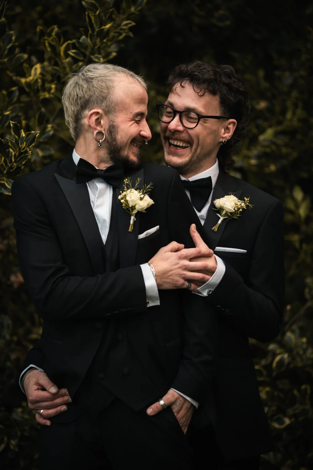 Two men in black tuxedos and bow ties, with white boutonnieres, sharing a joyful moment outdoors with greenery in the background.
