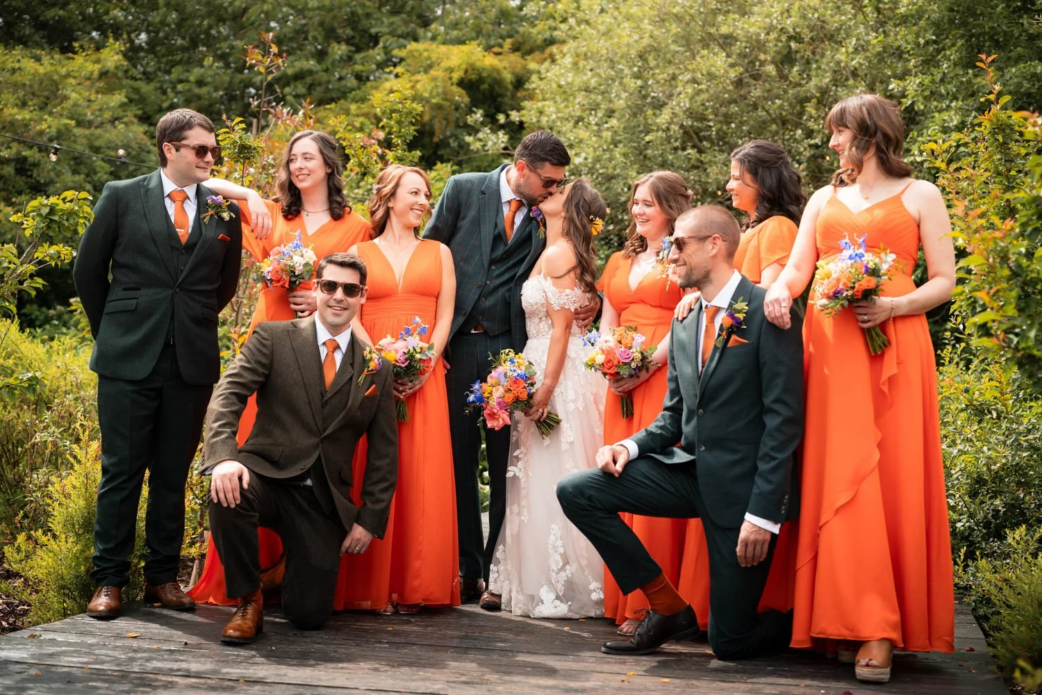 A wedding party outdoor photo with the bride and groom kissing surrounded by bridesmaids and groomsmen dressed in colorful attire, holding bouquets, on a wooden path surrounded by greenery.