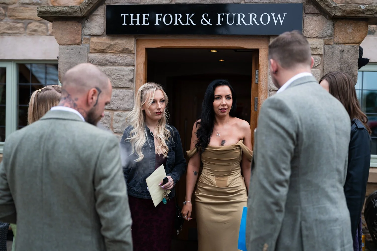 A group of people talking outside a building named 'The Fork & Furrow' with a woman in a strapless tan dress at the center.