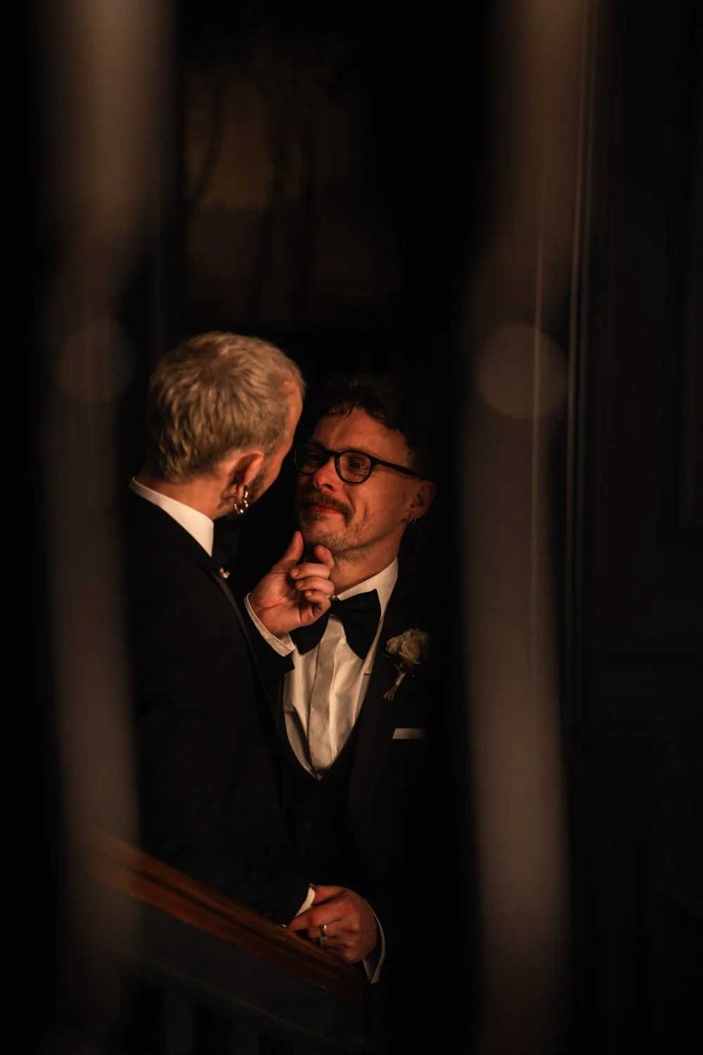 Two men in tuxedos sharing an intimate moment, one gently touching the other's face, in a dimly lit setting.