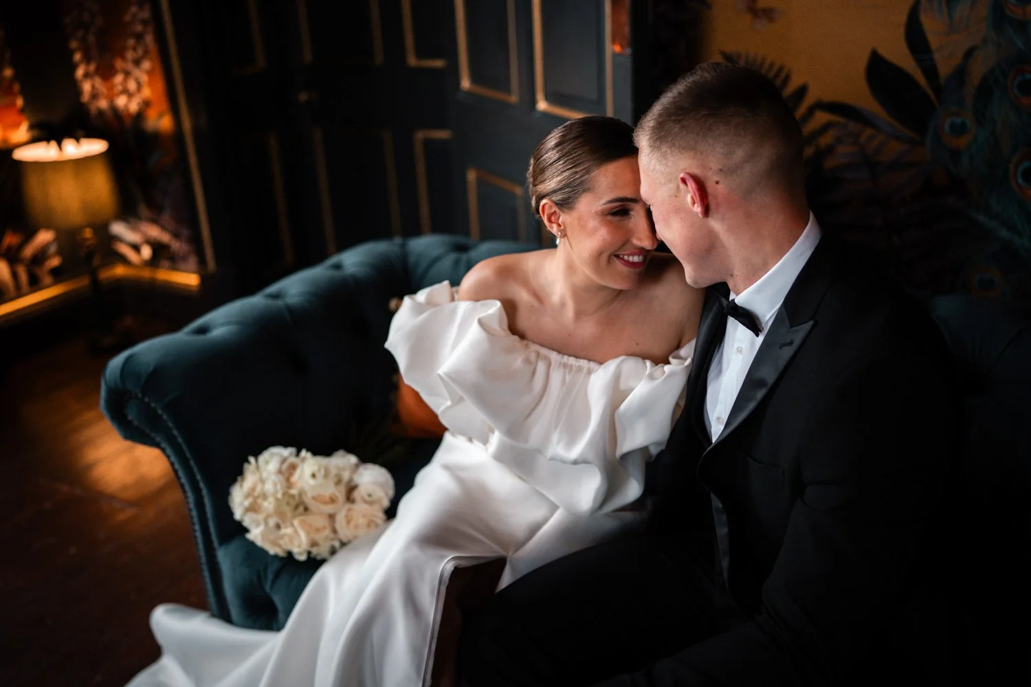 Bride and groom sitting close on a dark teal velvet sofa, smiling and touching foreheads, with a bouquet of white roses beside them, in a dimly lit room with dark walls and warm lighting.