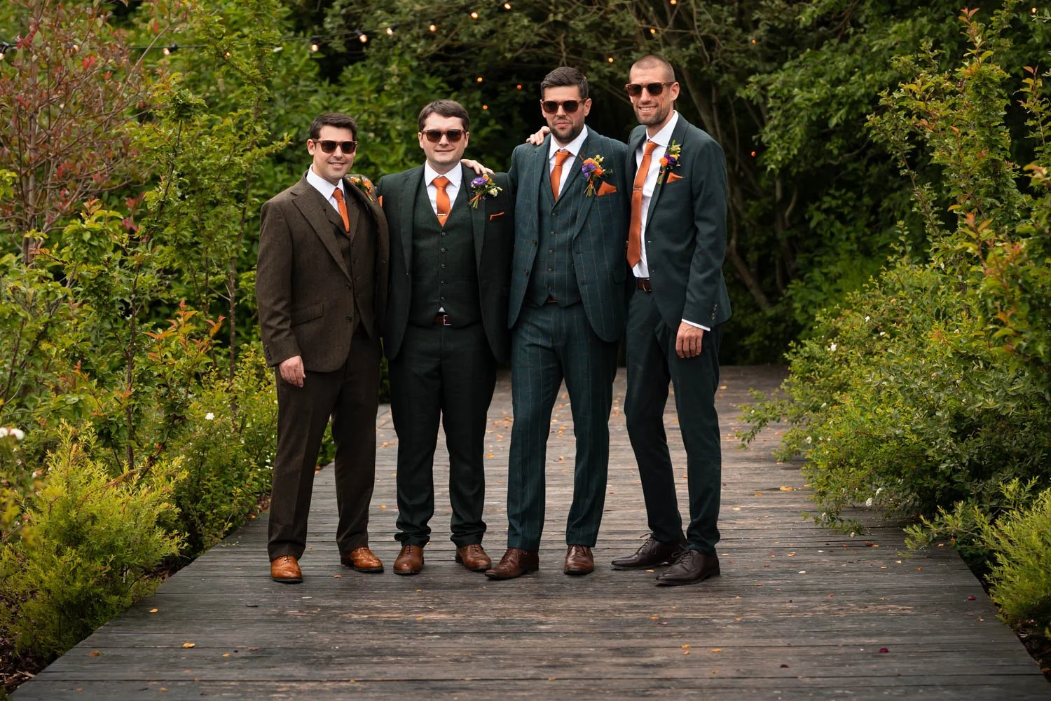 Five men dressed in suits and sunglasses standing on a wooden bridge outdoors, with greenery and string lights in the background.