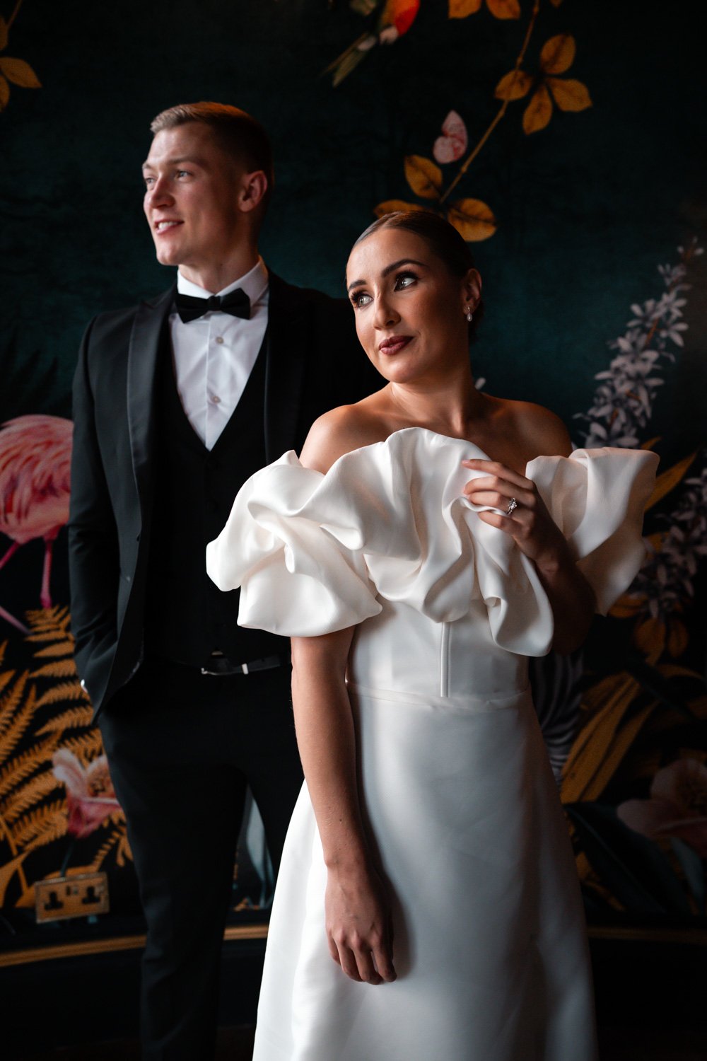 A bride and groom dressed in formal wedding attire standing against a dark floral background. The bride wears a white dress with voluminous ruffled sleeves and the groom wears a black tuxedo with a bow tie.