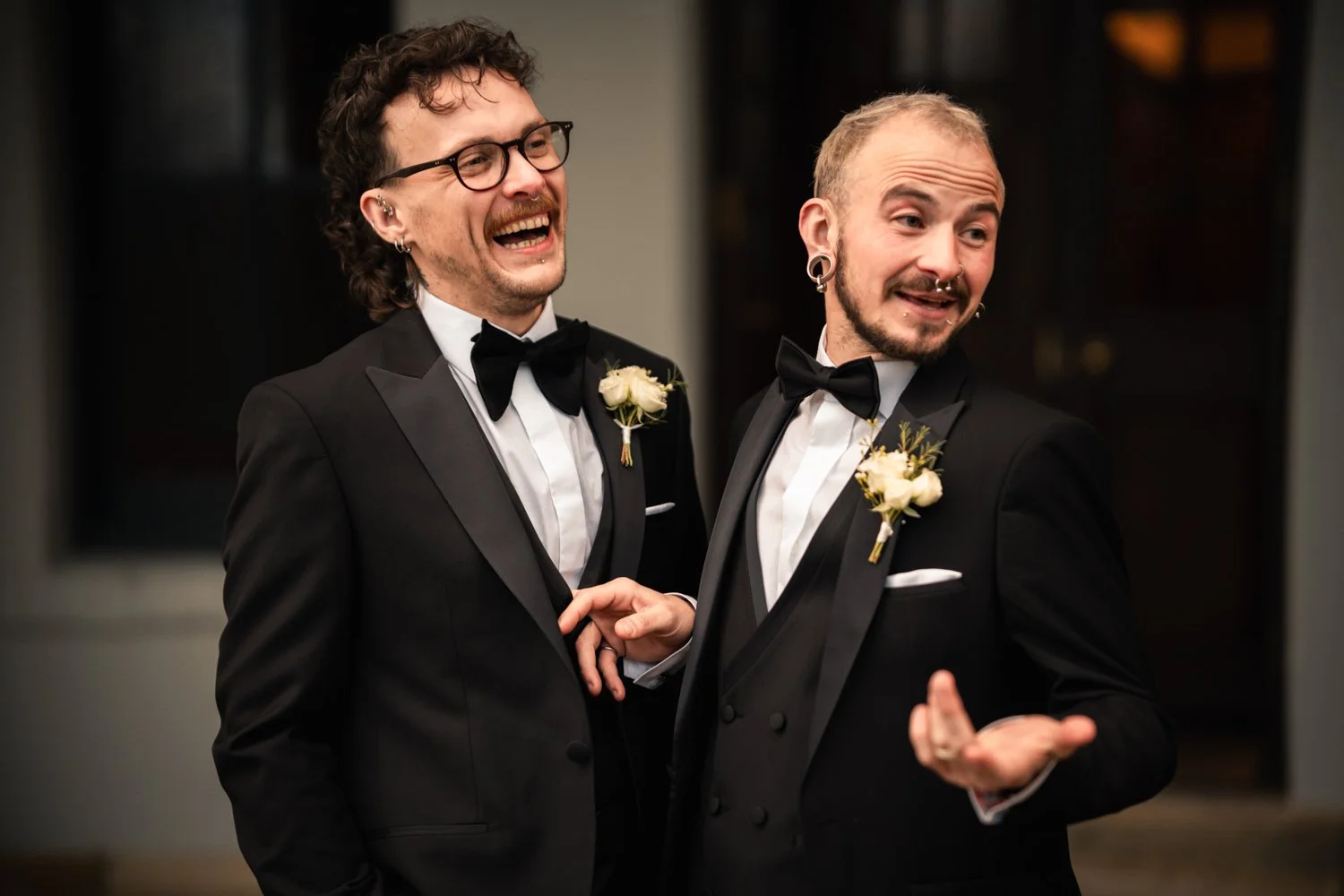 Two men in tuxedos with boutonnières, smiling and talking, at a formal event or wedding celebration.