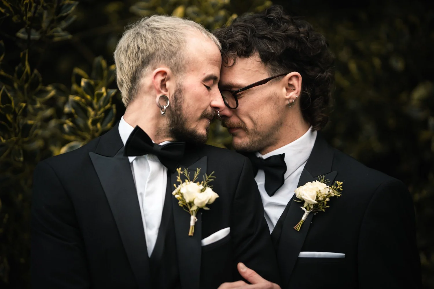 Two men dressed in black tuxedos with bow ties and white shirts, standing close with foreheads touching, in an outdoor setting. They each wear a white boutonniere and have pierced ears. One has blonde hair and a beard, the other has dark, curly hair 