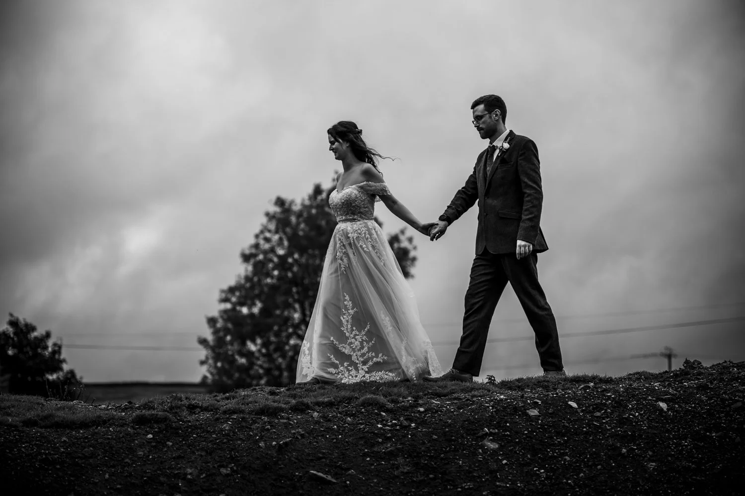 A black and white photo of a bride and groom holding hands, walking outdoors on a hilltop with a cloudy sky and trees in the background.