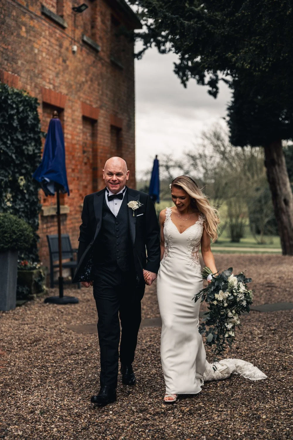 A bride and groom walking hand in hand outdoors on their wedding day, with the bride holding a large bouquet of white flowers and greenery, smiling.