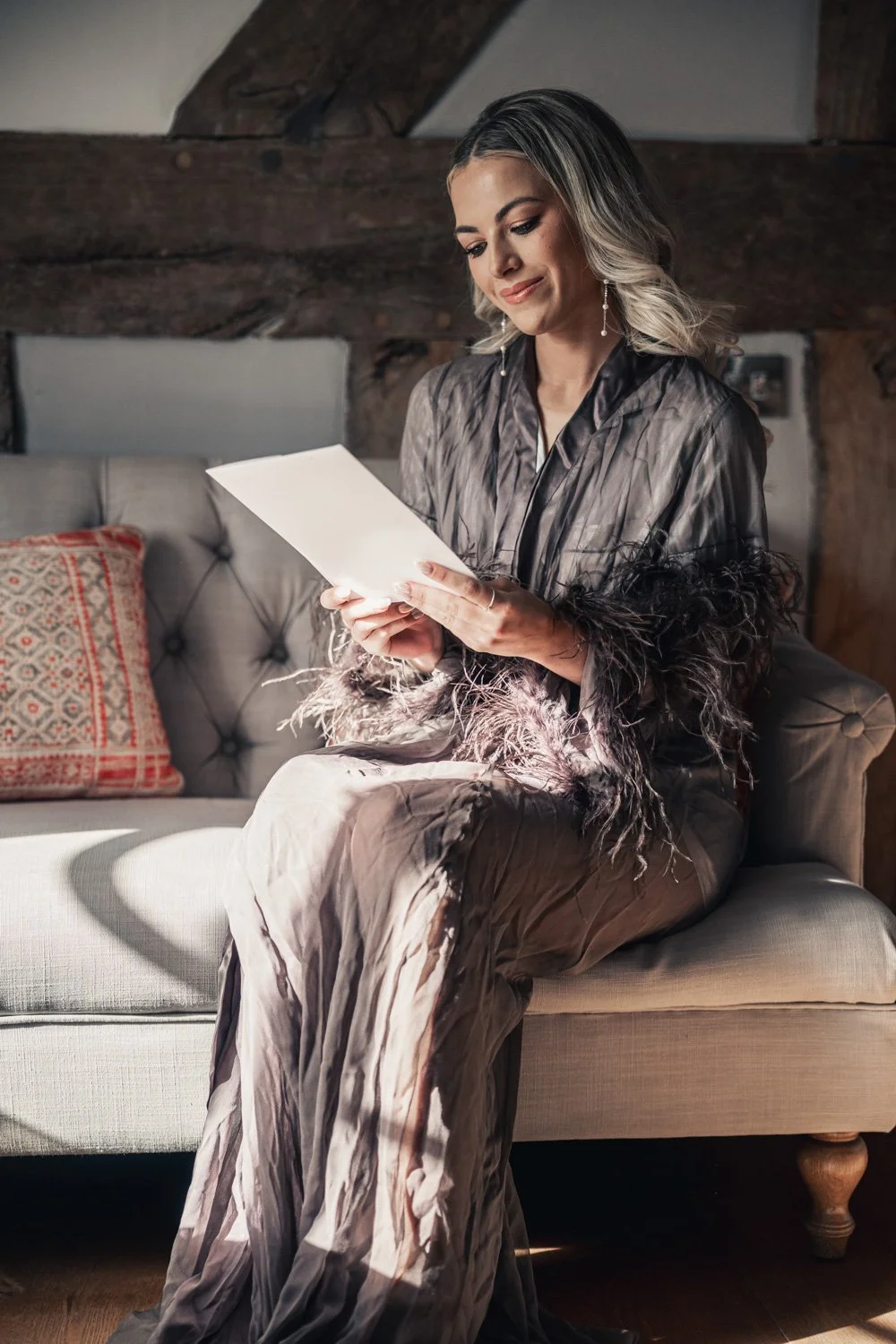 A woman sitting on a beige sofa in a cozy room with wooden beams, reading a script or letter, dressed in a satin blouse with feathered cuffs and matching pants.