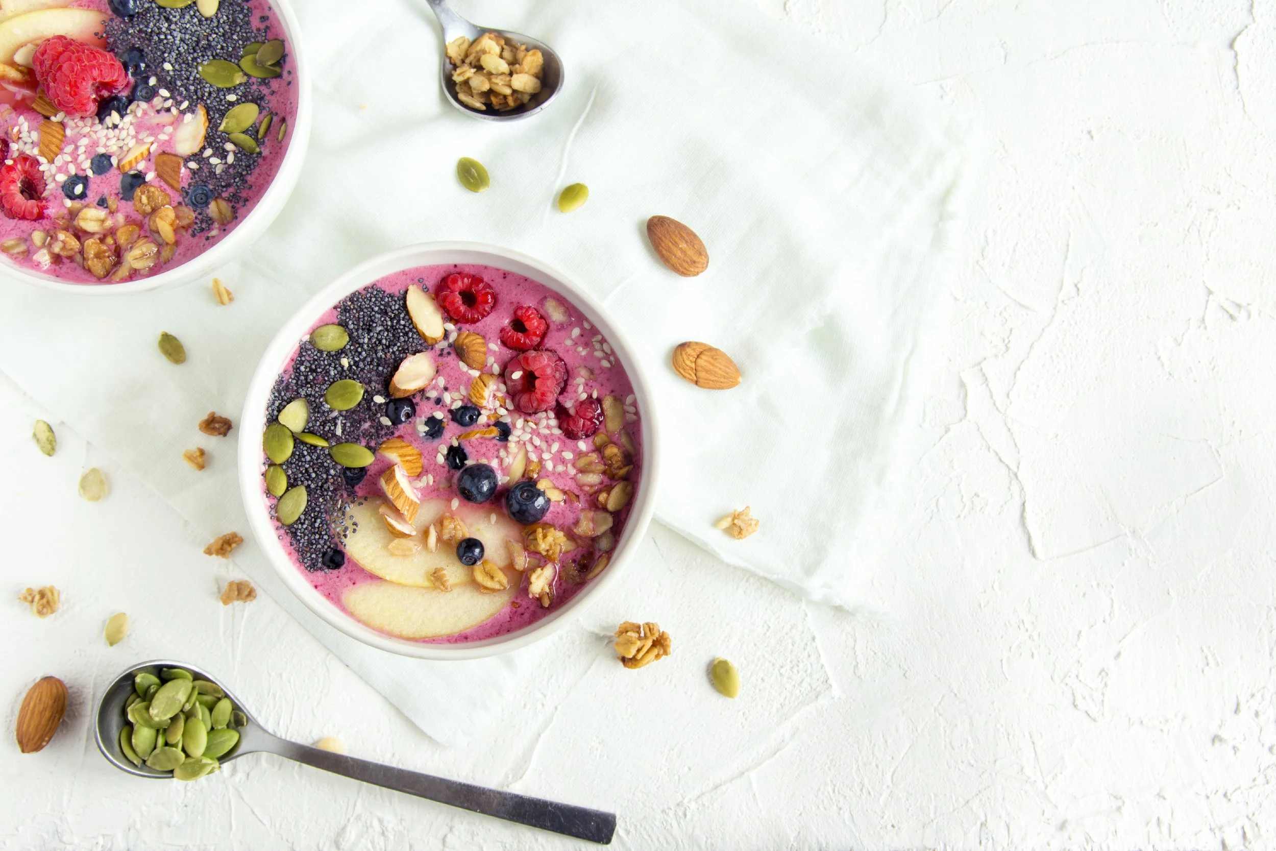 Two bowls of pink smoothie topped with mixed berries, banana slices, seeds, and nuts on a white textured surface with scattered nuts and seeds.