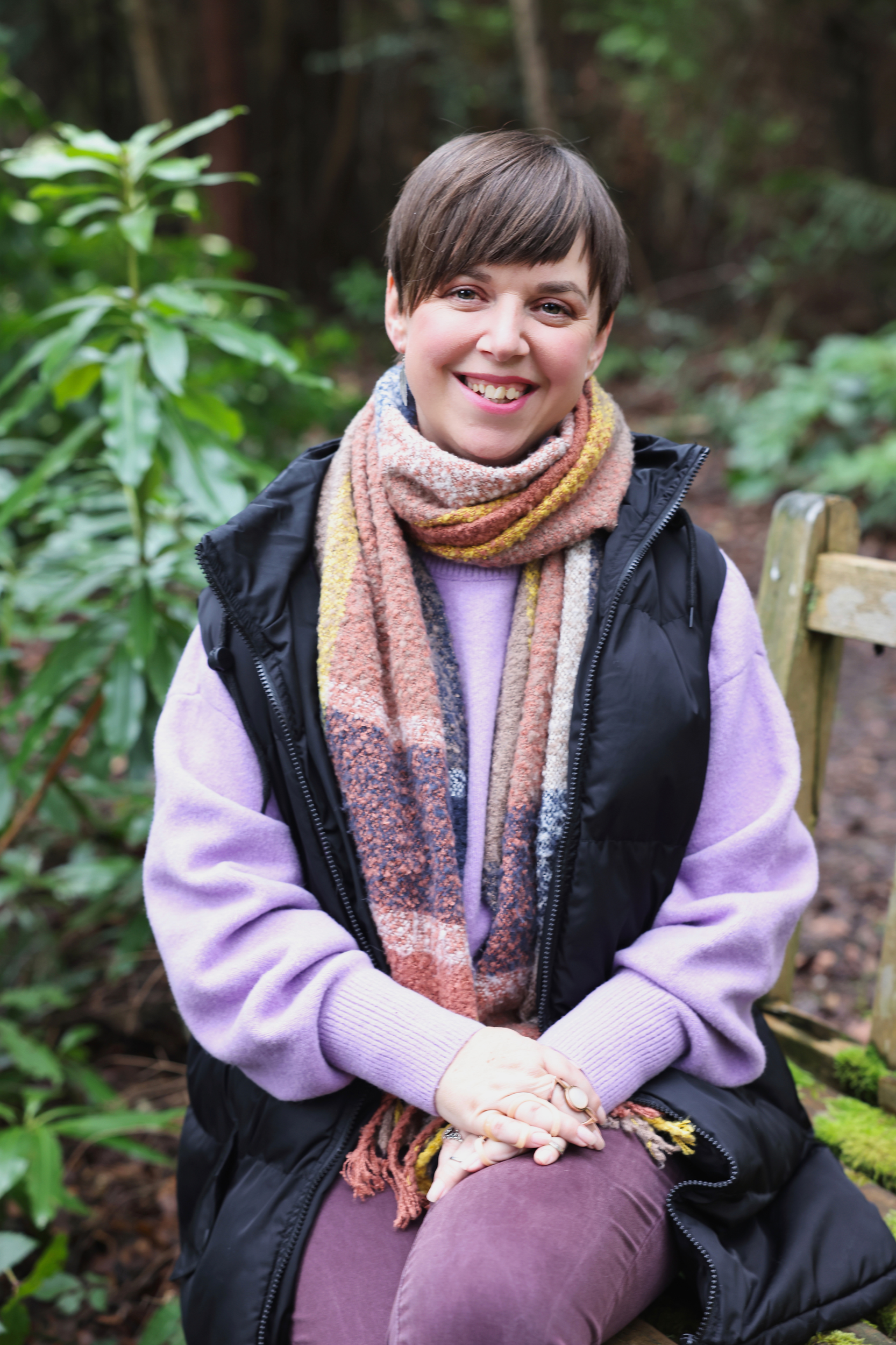 Lady, smiling, sat on a bench in the woodland, wearing purple jumper, scarf and jacket