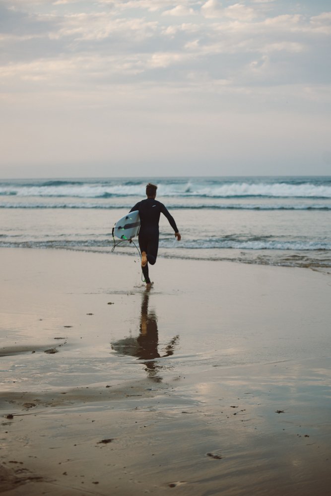 Person wearing a wetsuit carrying a surfboard running towards the ocean on a beach.