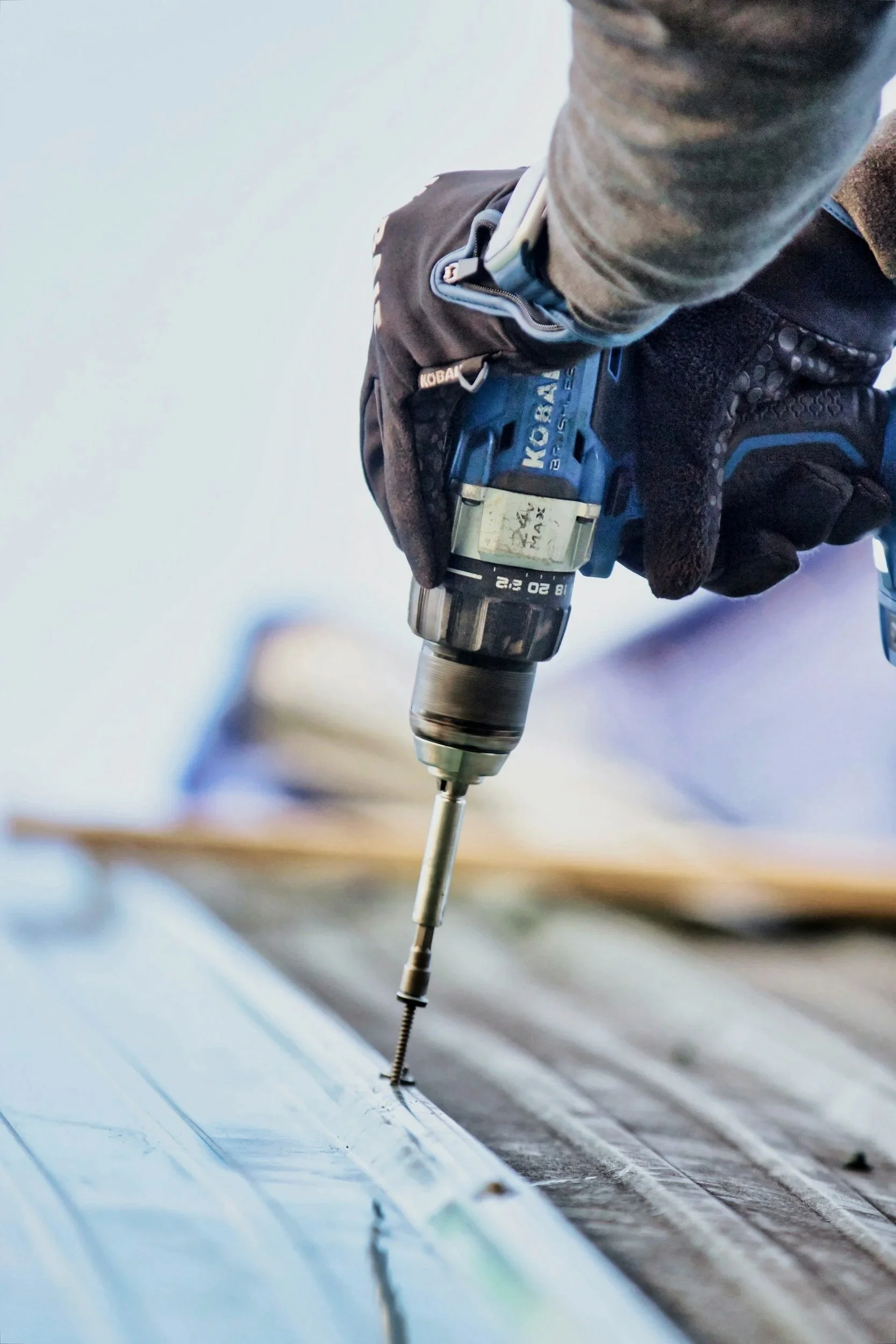 Close-up of a person using a cordless power drill to drill into a metal surface, wearing gloves and a gray long-sleeve shirt.