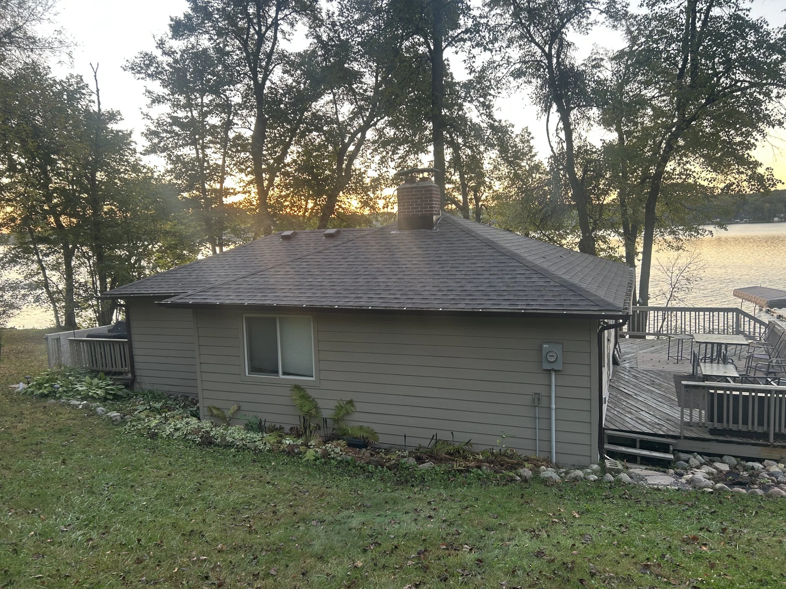 A house with gray siding, a shingled roof, and a brick chimney, surrounded by trees, with a deck featuring outdoor furniture and a view of a body of water at sunset.