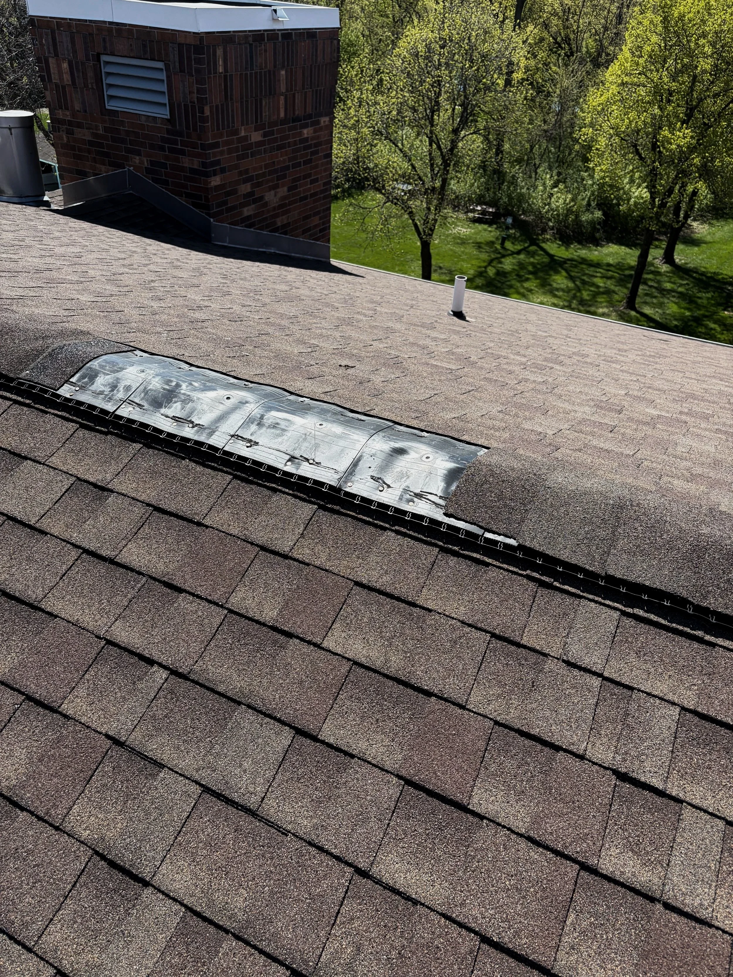 View from a rooftop, showing asphalt shingles, a damaged metal rain guard, and part of a brick chimney, with trees and green grass backyard in the background.