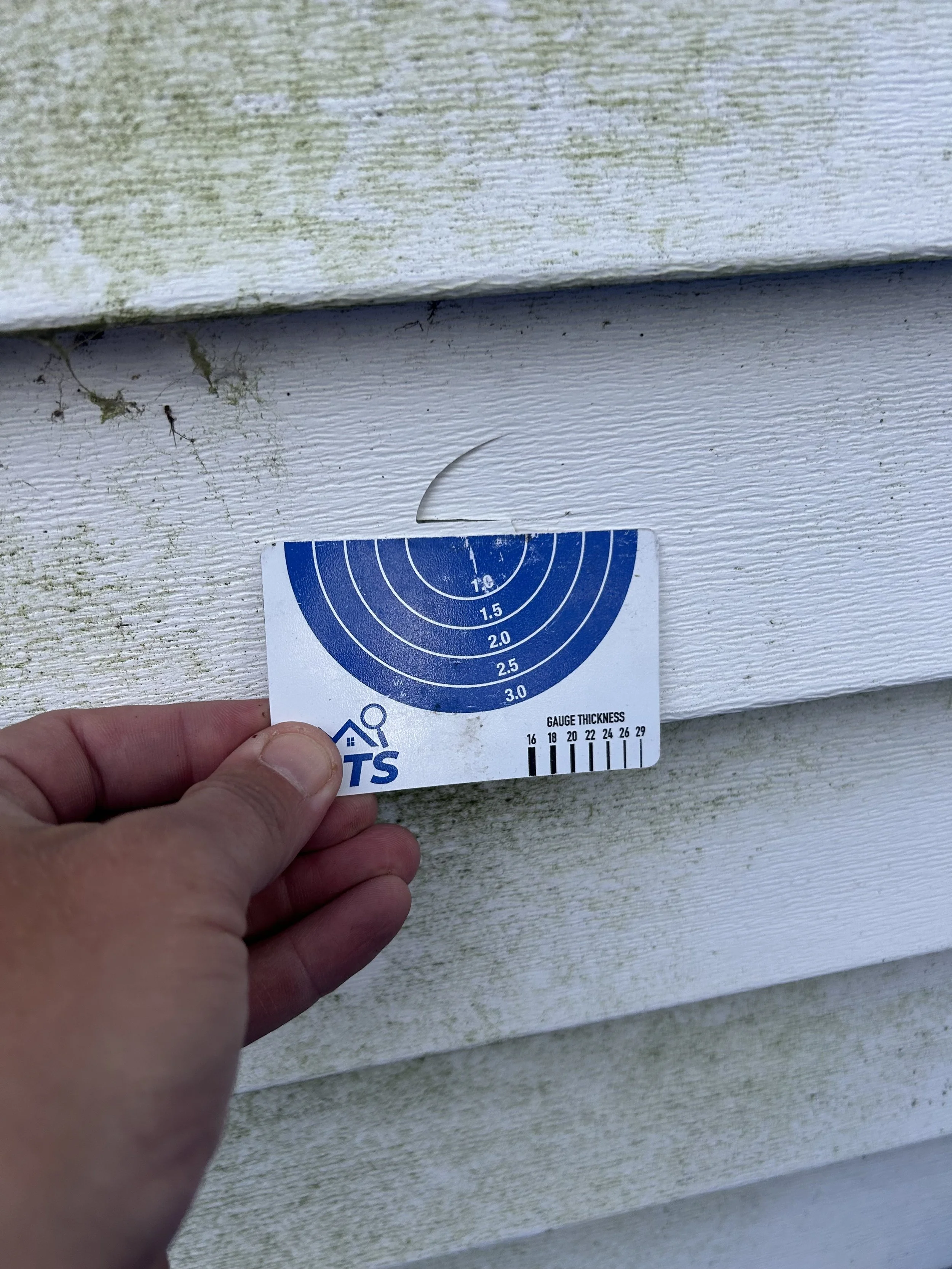 Person holding a gauge thickness measurement card against white plastic siding.
