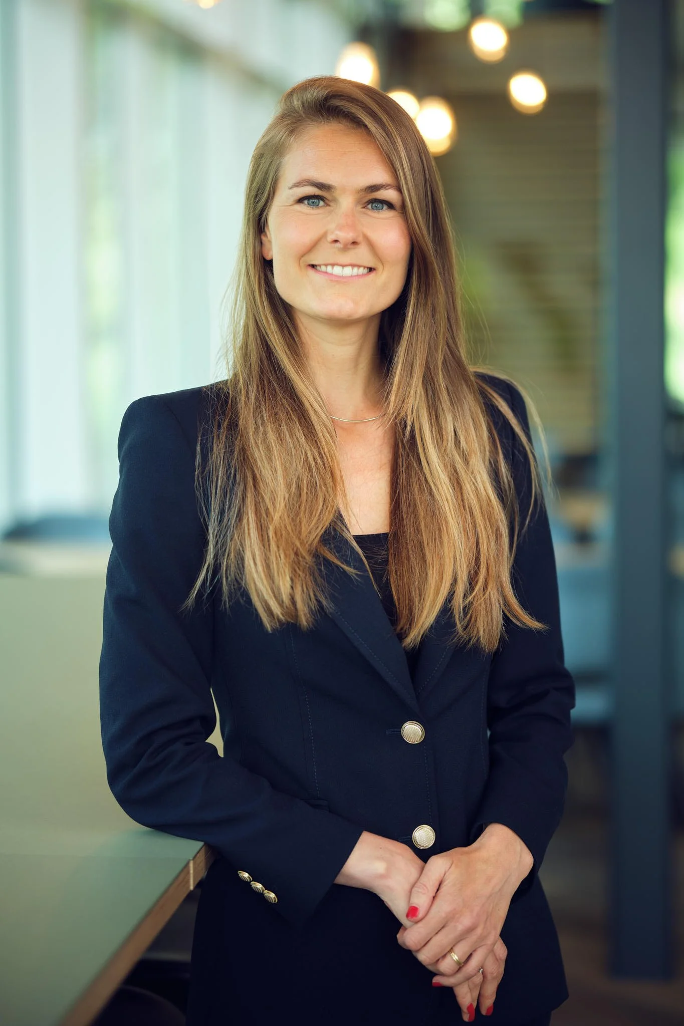 Woman with long dark blonde hair wearing a dark blue suit leaning on a table standing in an open light space with some windows and small lights in the background