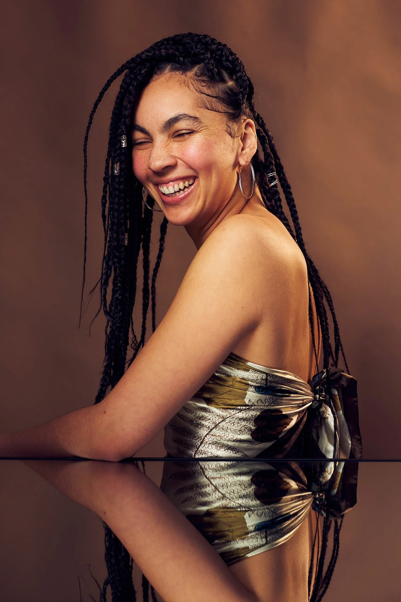 A woman with long black braided hair laughing with her eyes closed posing above a mirror against a beige background