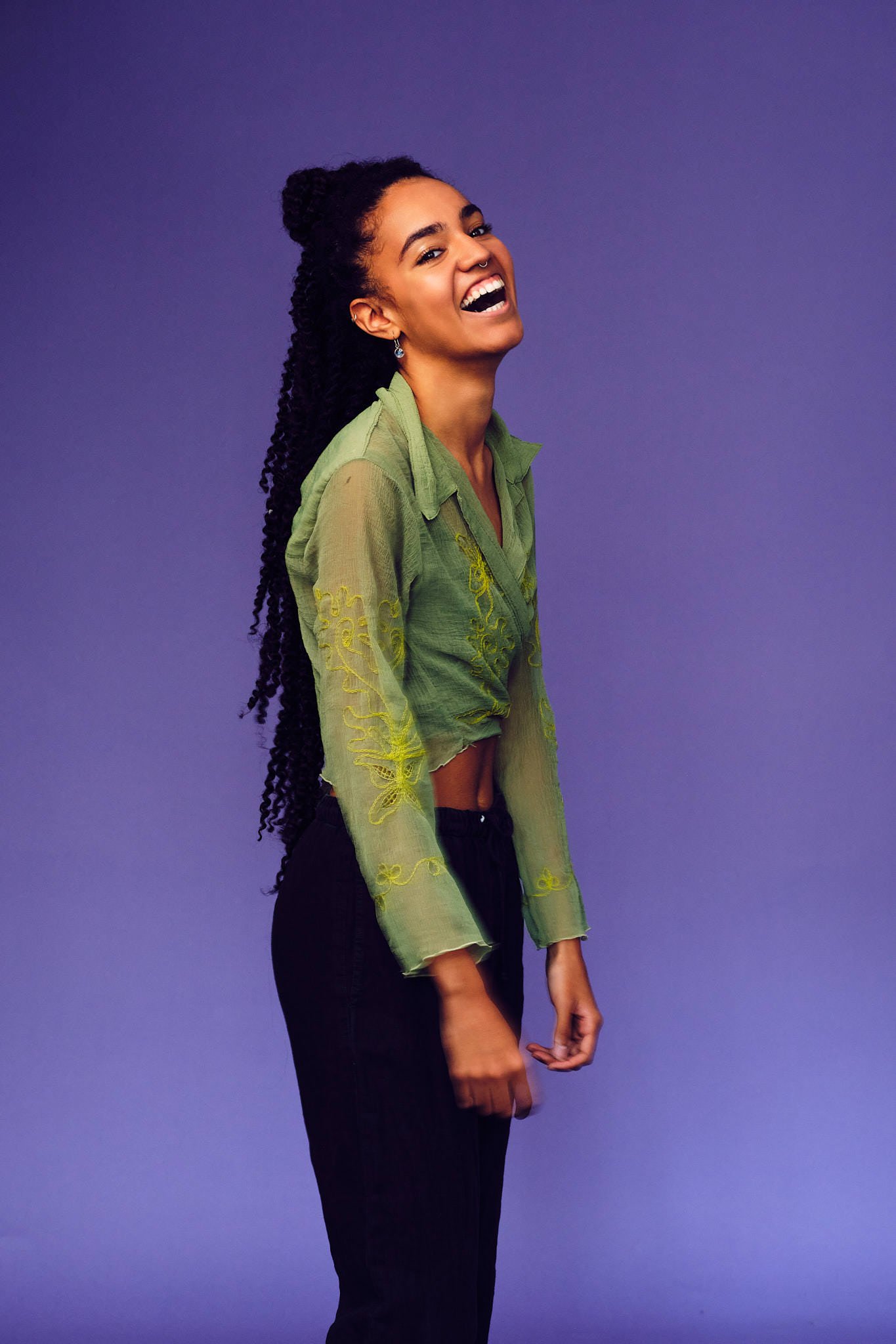 Young woman with long black braids, wearing a green long sleeve top and black trousers, is laughing widely, against a purple background.