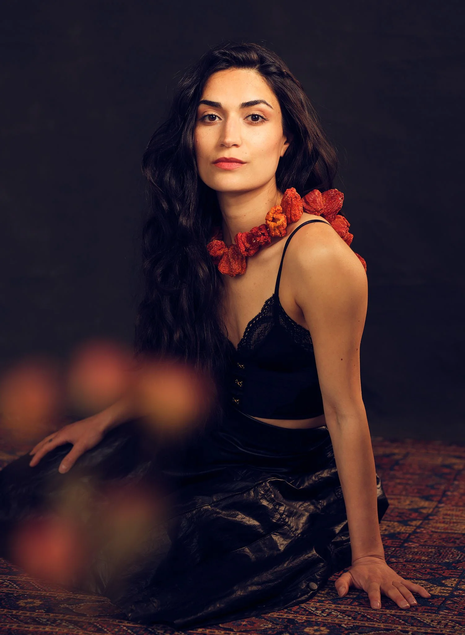Woman dressed in a black top, shiny black skirt and garland of red bell peppers sitting on a patterned floor rug against a dark background