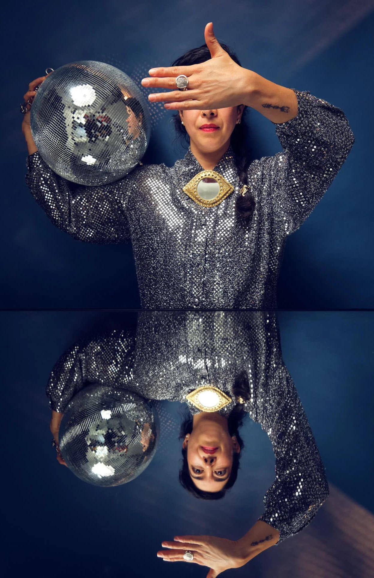 A woman with long black hair in one side braid wearing a silver sequined shirt and eye shaped mirror at her neck with one hand in front of her face posing with a disco ball above a mirror against a dark blue background
