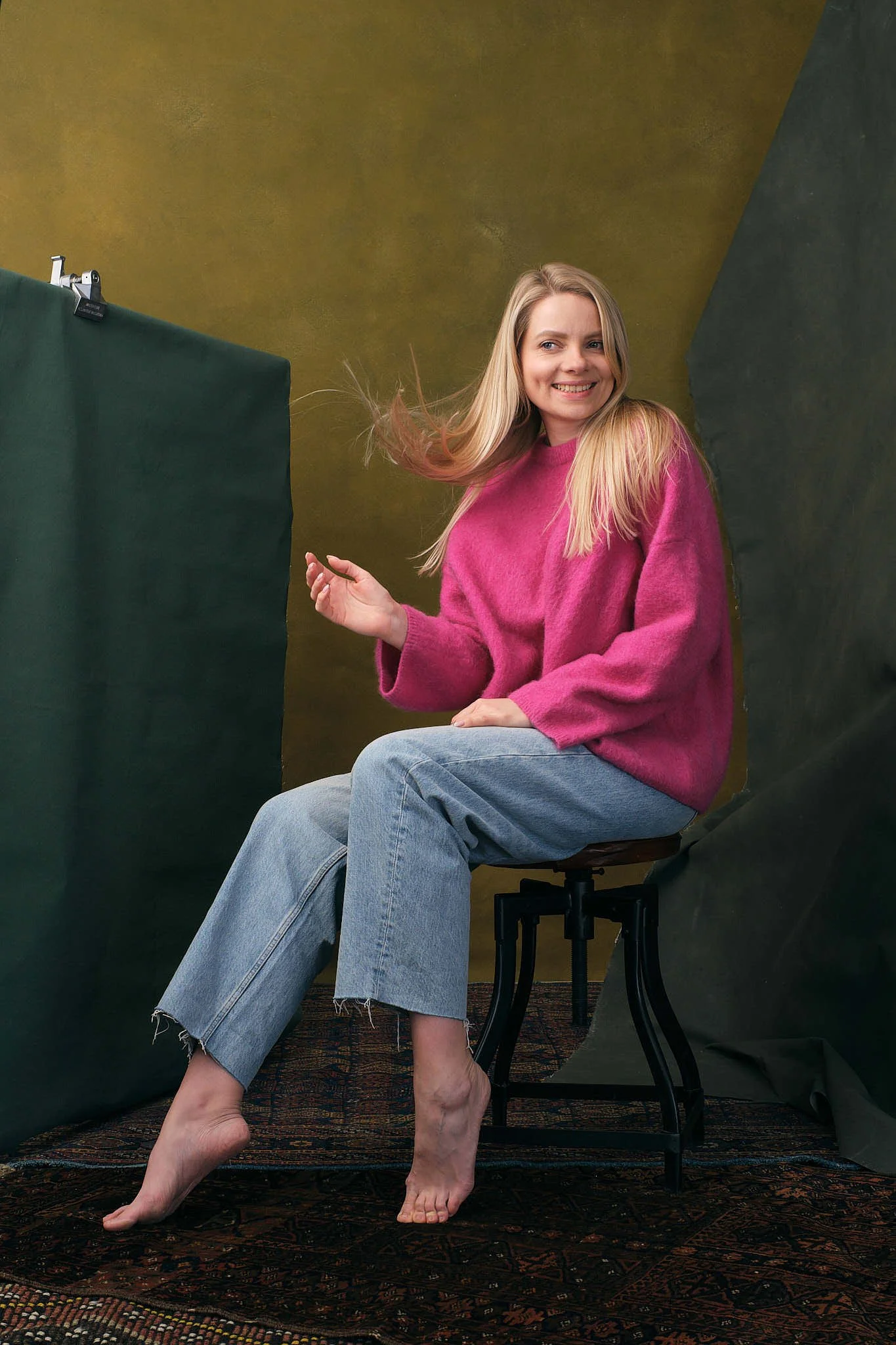 A woman with long blonde hair wearing a pink sweater and jeans is sitting barefoot on a stool, smiling with her hair flying to the side, against a multicoloured backdrop.