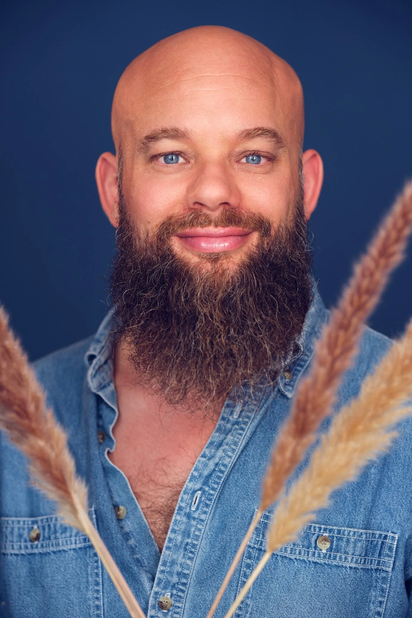 A smiling man with a beard and blue eyes wearing a denim shirt, with pampas grass in the foreground and a dark blue background.