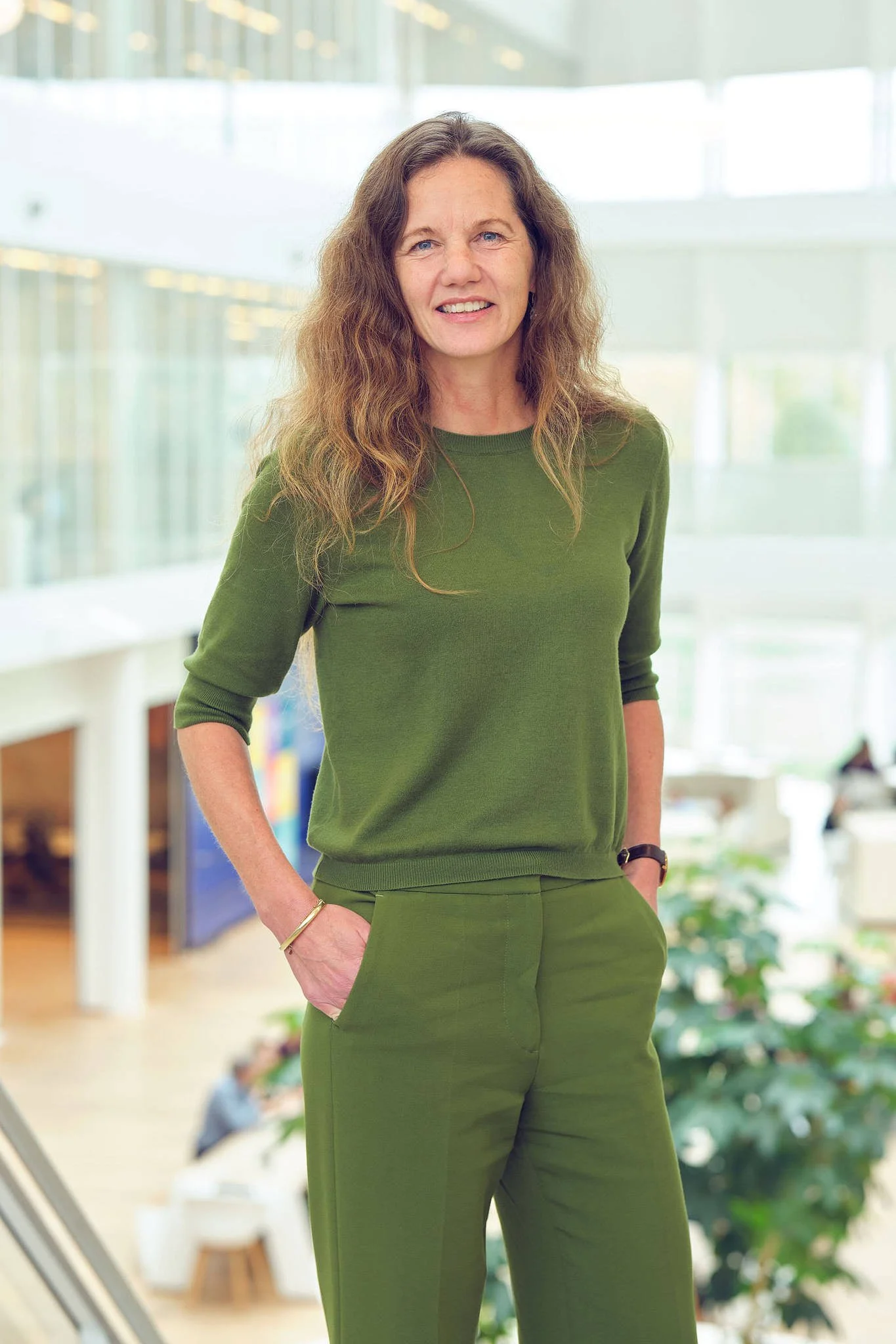 A woman standing in a modern indoor space, wearing a green long-sleeve top and matching green trousers, smiling and looking at the camera.