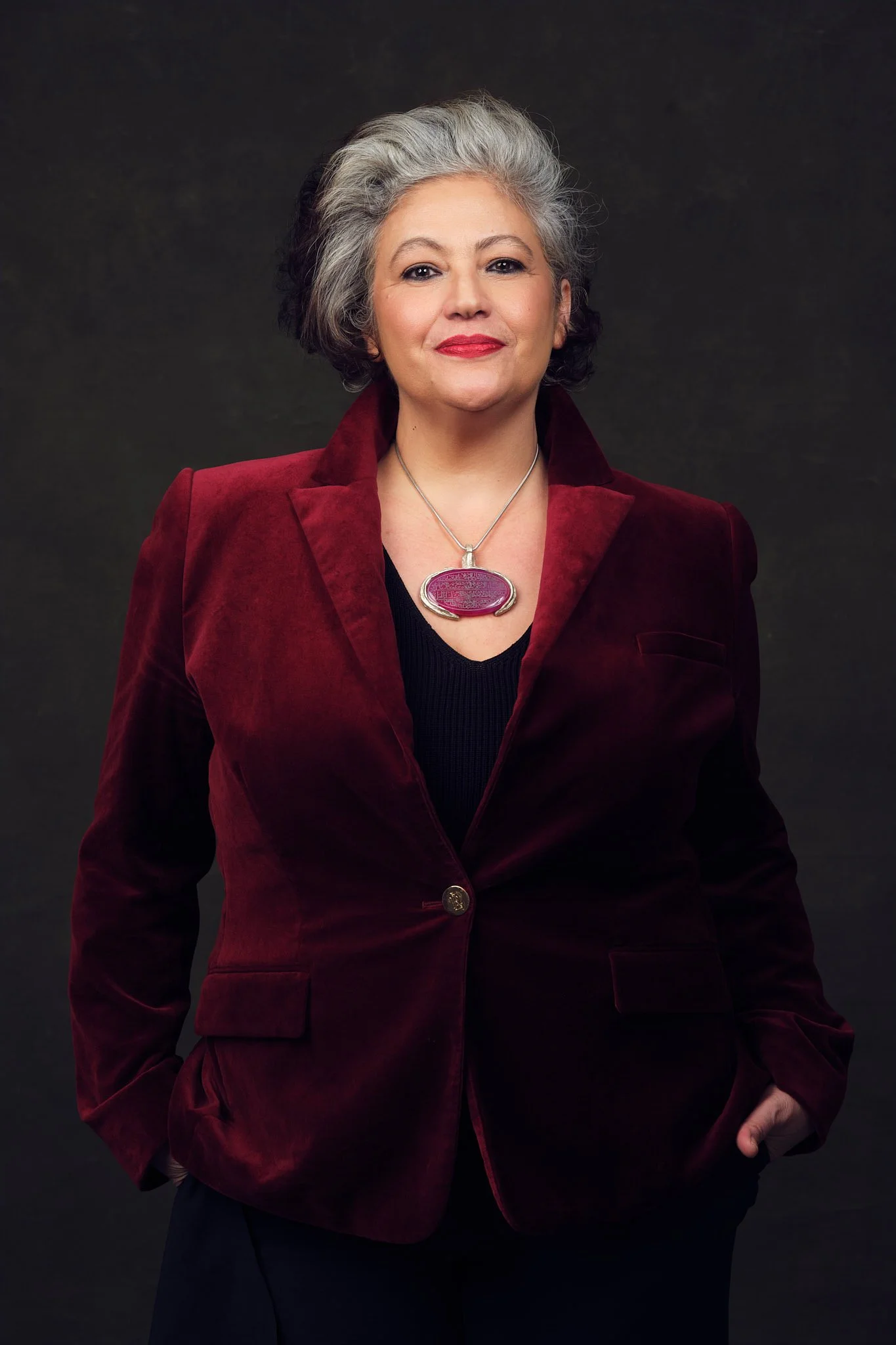 Woman with grey and dark hair wearing a burgundy red jacket and red pendant posing confidently against a dark background