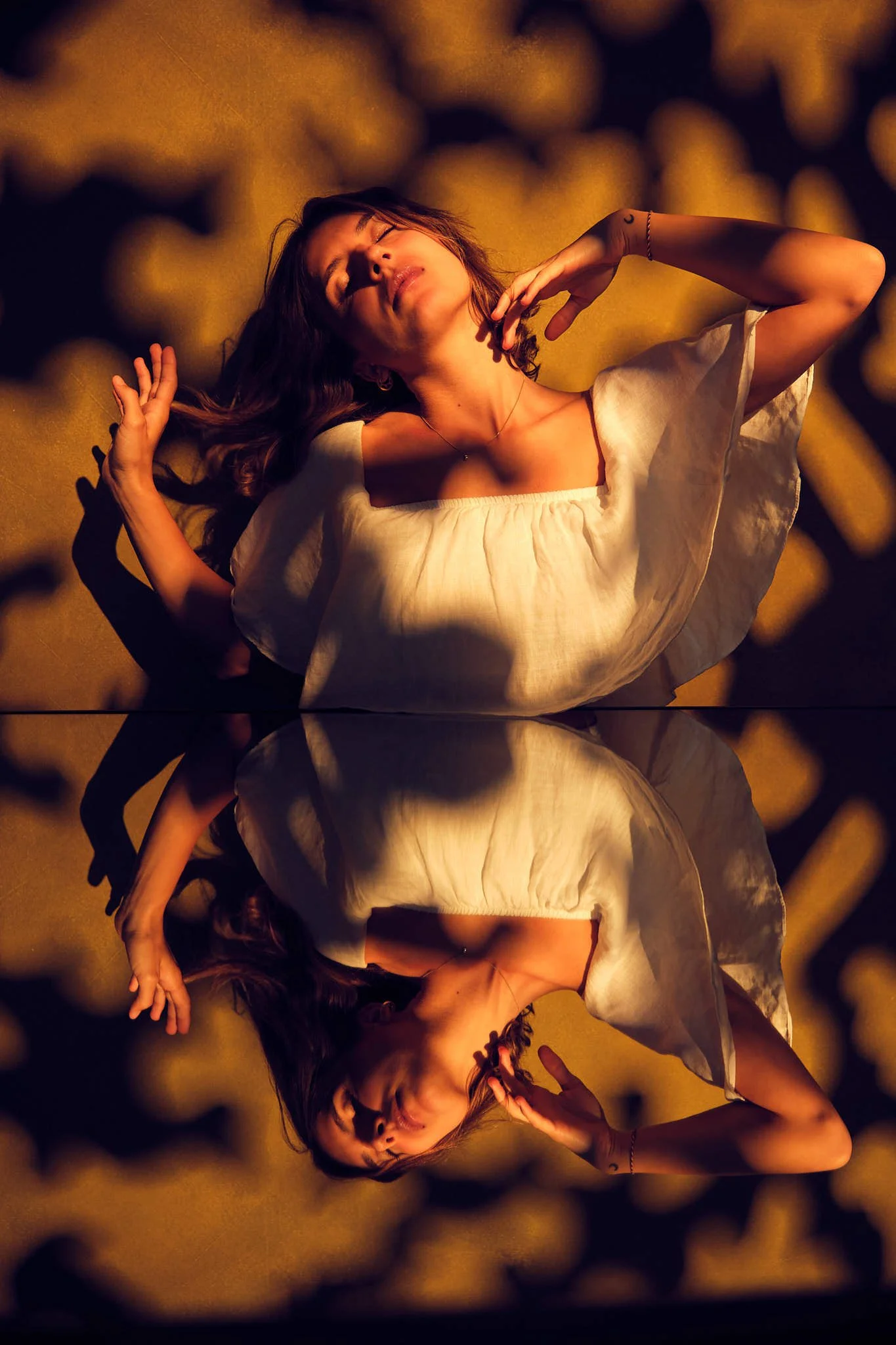 A woman wearing a white top dancing with her eyes closed and hands raised above a mirror against a dappled dark yellow background