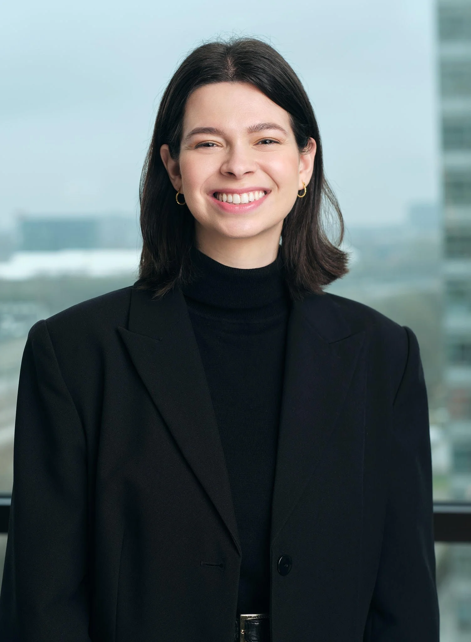 A woman with shoulder-length dark hair, wearing a black blazer and a black turtleneck, smiling in front of a window with city buildings in the background.