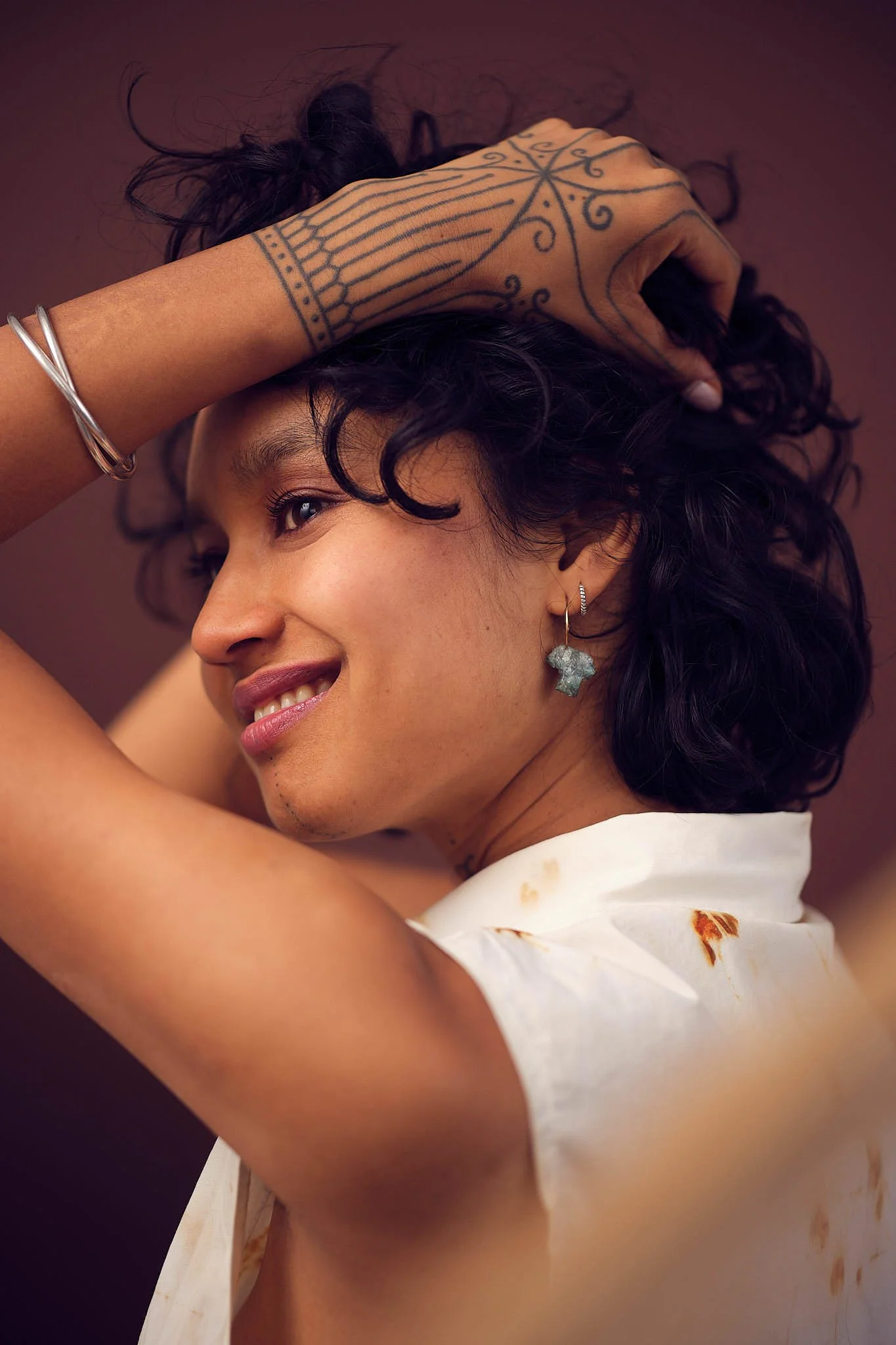 A woman with curly dark hair holding her head with one hand, smiling, wearing earrings, a silver bracelet, and a sleeveless white top with dried flowers embedded in it, against a dark brown background.