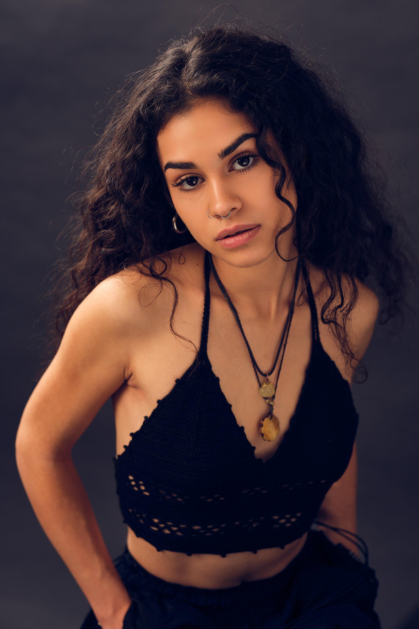A young woman with curly dark hair and light brown skin posing against a dark background. She is wearing a black crochet halter top and necklaces, with hoop earrings and a nose ring. She has a neutral expression and her head is slightly tilted.