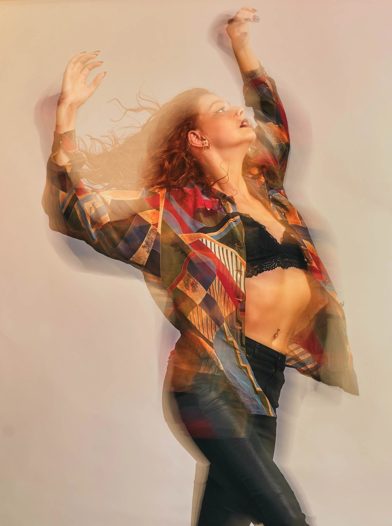 A woman with curly red hair wearing a colorful patterned jacket and black lace bralette posing against a plain wall, with motion blur effect.