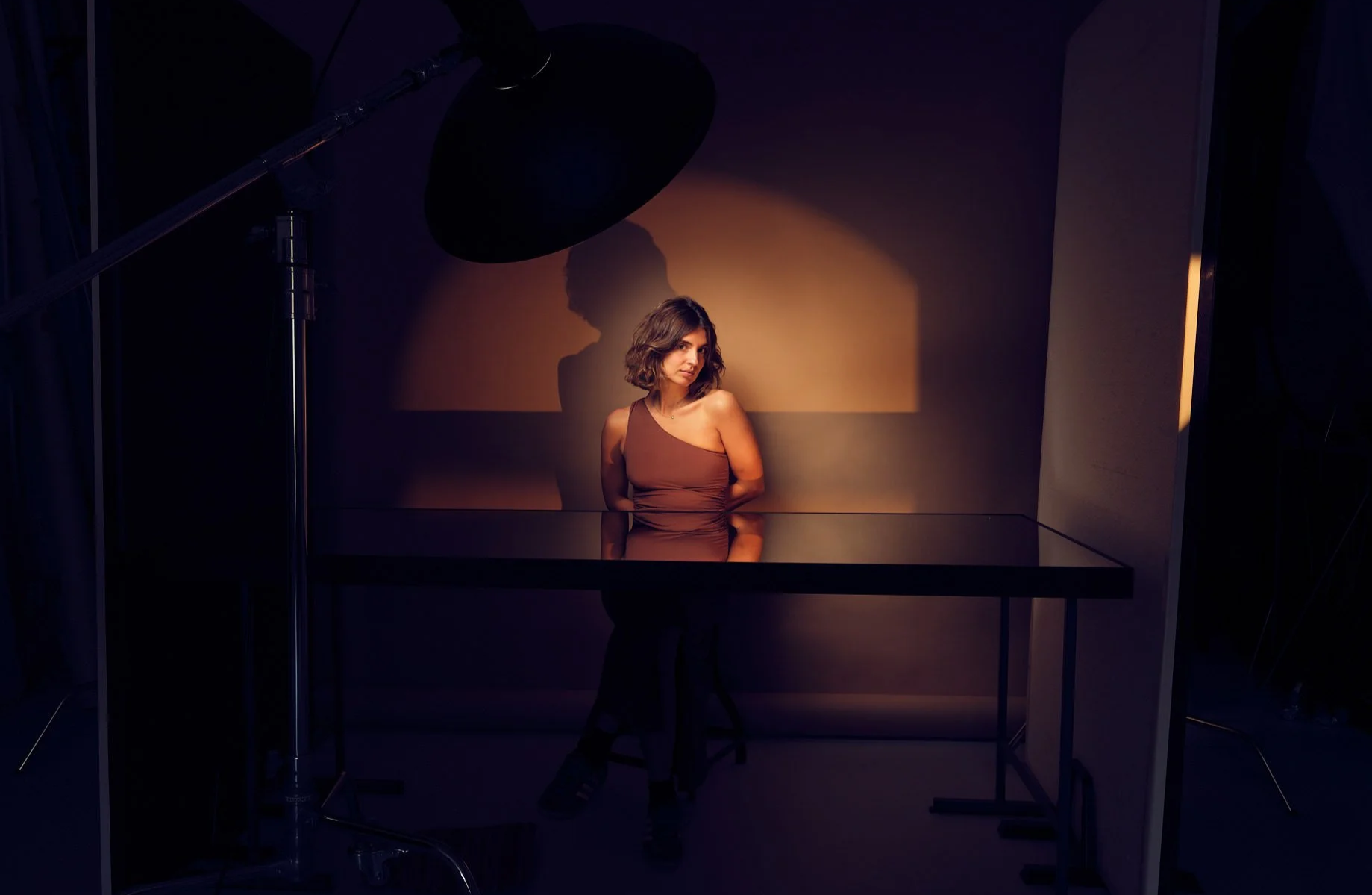 A woman wearing a one-shoulder brown top with her hands behind her back posing above a mirror casts a shadow against a dark beige background