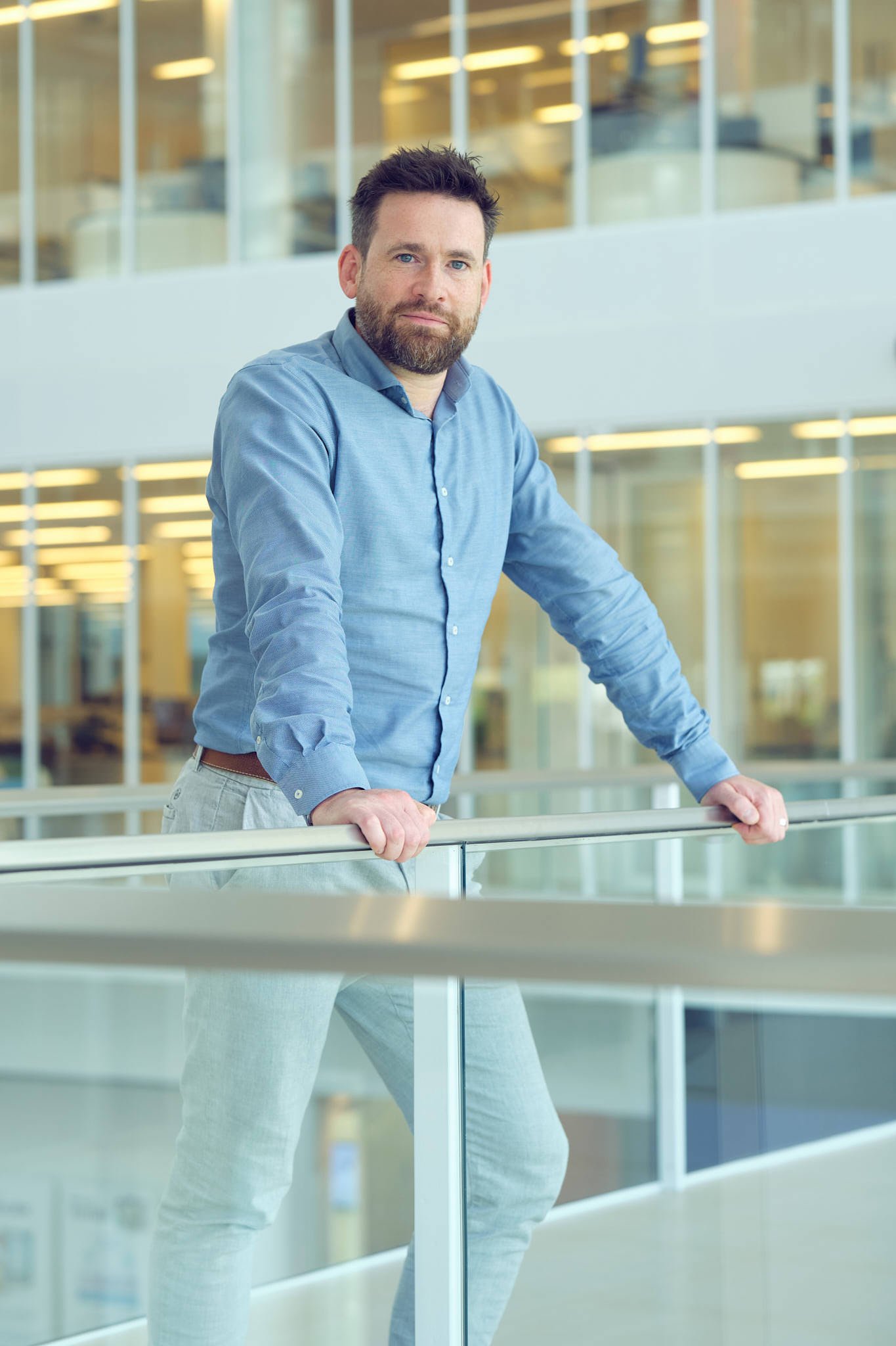 A man with a beard and blue shirt leaning on a railing inside a modern office building with glass walls and bright lighting.